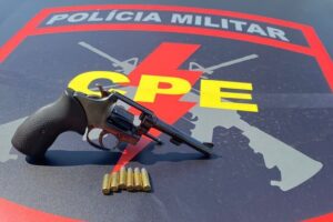 Revólver com munição em cima de um capô de viatura com logo do CPE da Polícia Militar