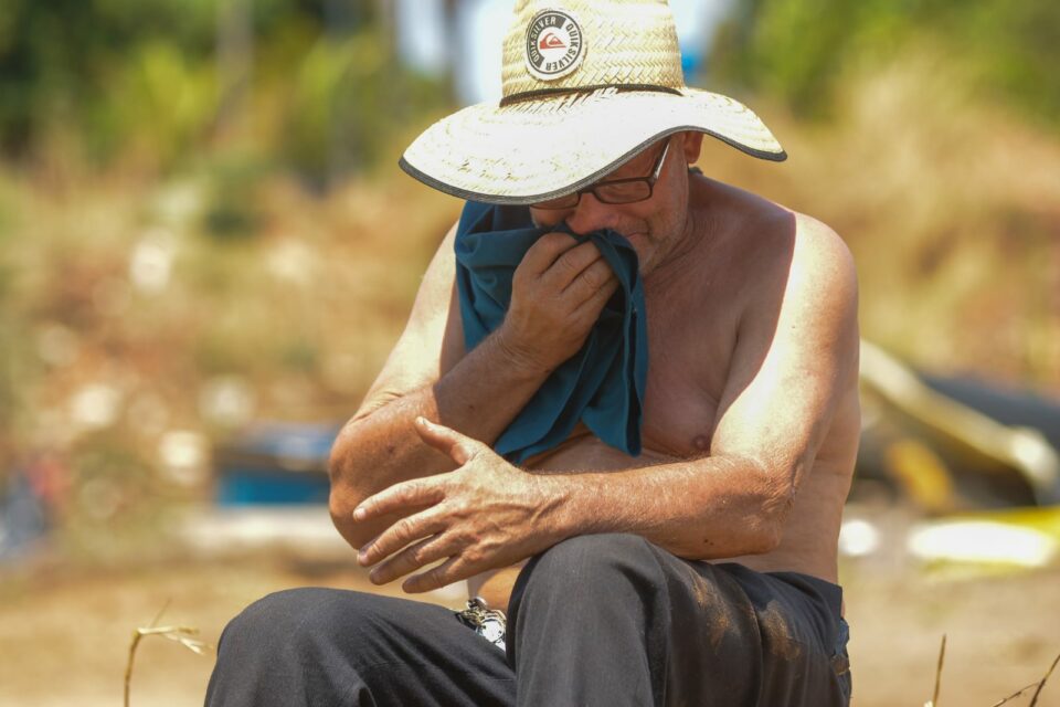 Homem limpa a boca com camiseta enquanto está sentado ao sol com cara de choro