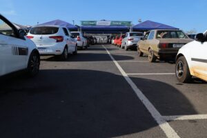 Fila de carros de dois lados em estacionamento. Ao fundo cartaz de vacinação em Aparecida de Goiânia