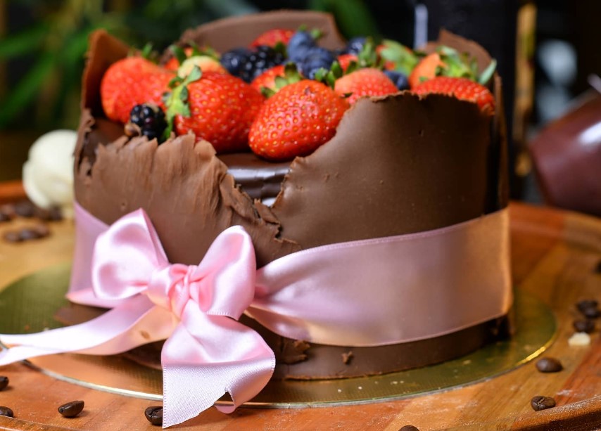 bolo de frutas vermelhas envolto em uma casca de chocolate, finalizado com um laço ao redor. A receita é opção da Confeitaria Vitória, opção entre as confeitarias em Aparecida de Goiânia